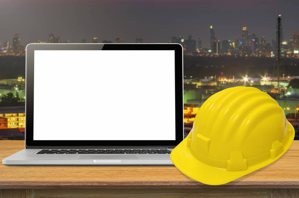 Casque de sécurité jaune et ordinateur portable sur le bureau avec arrière-plan de l'industrie, blanc à l'écran
. - Photo, image