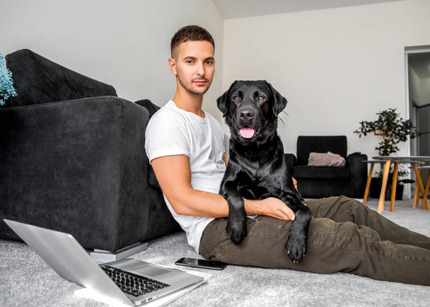 freelancer guy sitting at home working with a dog in an embrace, black labrador. - Photo, Image