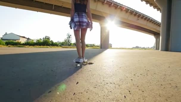 Hermosa chica monta un monopatín bajo el puente en el sol, en el marco de la pierna
 - Imágenes, Vídeo