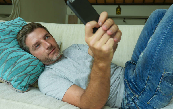 lifestyle indoors portrait of young happy and attractive man at home sofa couch using internet social media app on mobile phone networking relaxed and cheerful from modern apartment - 写真・画像