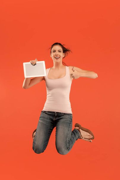 Image of young woman over blue background using laptop computer or tablet gadget while jumping. - Foto, Imagem