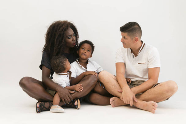 Mezclador de raza familiar sentado en el suelo con 2 niños. Sonriente pareja sentada con dos hijos aislados
. - Foto, imagen