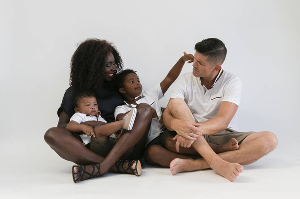 Mezclador de raza familiar sentado en el suelo con 2 niños. Pareja sonriente sentada con dos hijos aislados. Padre habla con el niño
. - Foto, imagen