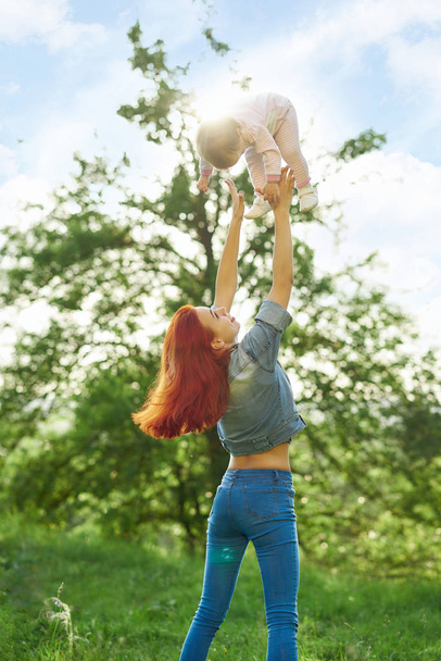 Young mother throwing little daughter up in the air in green summer garden. - Foto, afbeelding