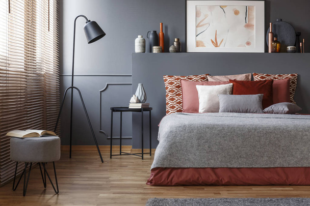 Stool and lamp near bed with pink and grey bedding in bedroom interior with poster on headboard - Foto, afbeelding