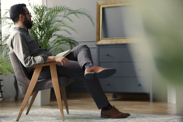 Low angle of hipster sitting on wooden armchair in grey vintage interior with plant - Foto, Imagem