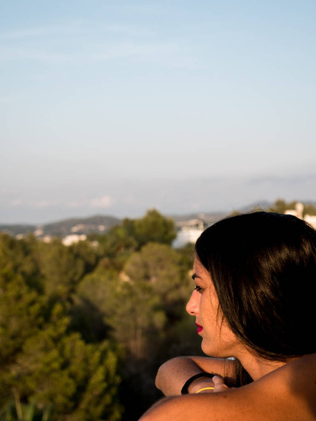 Красива молода жінка в профілі дивиться з балкона на горизонті з великою кількістю рослинності навколо неї
 - Фото, зображення