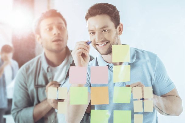 smiling young businessman writing on sticky notes while shocked colleague standing behind  - Photo, Image