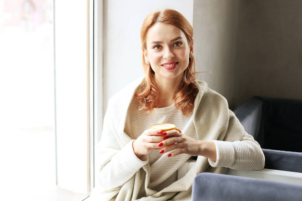 Beautiful redhead woman, sitting near coffee shop window holding yellow cup of hot beverage, wearing winter autumn season white knitted sweater & big loose scarf on shoulders. Background, close up. - Фото, зображення