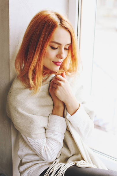 Beautiful redhead young woman with red lipstick on lips sitting by the window of coffee shop, wearing winter autumn season white knitted sweater & big loose scarf on shoulders. Background, close up. - Photo, Image