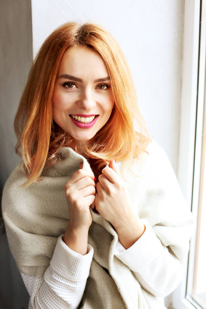 Beautiful redhead young woman with red lipstick on lips sitting by the window of coffee shop, wearing winter autumn season white knitted sweater & big loose scarf on shoulders. Background, close up. - Φωτογραφία, εικόνα