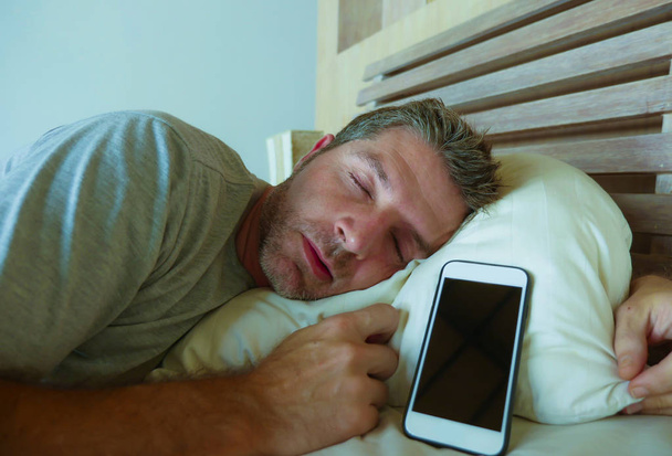 lifestyle indoors portrait of young internet addict man sleeping on bed holding mobile phone in his hand in smartphone and social media overuse and online addiction and obsession concept - Photo, Image