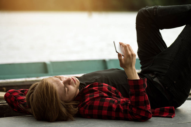 Young man in plaid shirt with long hair is lying down on dock and using phone. - 写真・画像