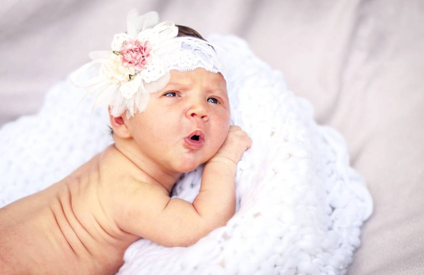 A sweet Caucasian newborn baby girl with a surprised look on a white pillow with flower hair bow on her head. A cute two week old infant portrait. - Photo, Image