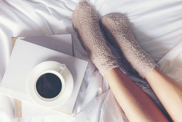Sulje jalat naiset valkoisella sängyllä. Naiset lukevat kirjaa ja juovat kahvia aamulla rentoutua tuulella talvikaudella. Elämäntapakäsite
. - Valokuva, kuva