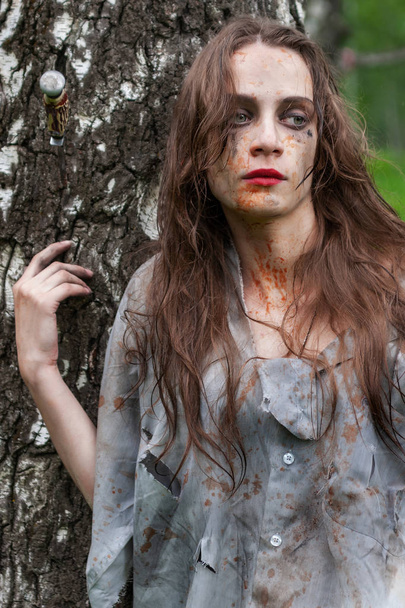 Beautiful young dirty mad and manic looking girl wearing torn clothes and smeared with mud and dried blood stands and huddles to a tree in the forest. Copy space. Concept design. - Foto, afbeelding