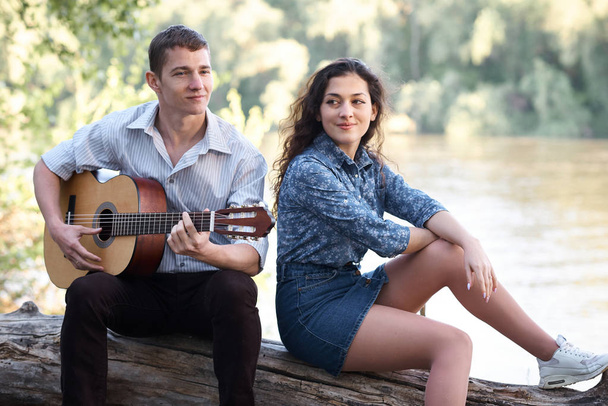 νεαρό ζευγάρι κάθεται σε ένα αρχείο καταγραφής από το ποτάμι και να παίζει κιθάρα, καλοκαίρι φύση, έντονο ηλιακό φως, σκιές και πράσινα φύλλα, ρομαντικά συναισθήματα - Φωτογραφία, εικόνα