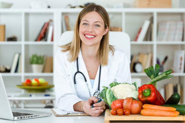 Portrait of beautiful smiling nutritionist looking at camera and showing healthy vegetables in the consultation. - Zdjęcie, obraz