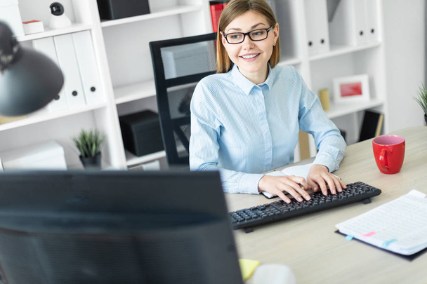 Una joven con gafas se sienta en una mesa en la oficina, sostiene un lápiz en la mano y escribe el texto en el teclado
. - Foto, imagen