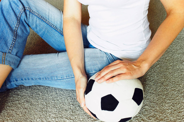 A woman in blue jeans close-up sits on a beige carpet and holds a soccer football ball - Fotoğraf, Görsel