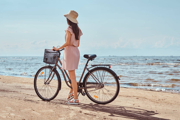 Back view of a brunette girl dressed in dress and hat walking with bicycle on the beach on a sunny day. - Photo, Image