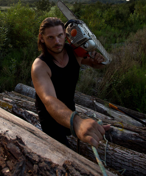 Young strong man lumberjack carrying chainsaw on shoulder and climbing on pile of logs - Fotoğraf, Görsel