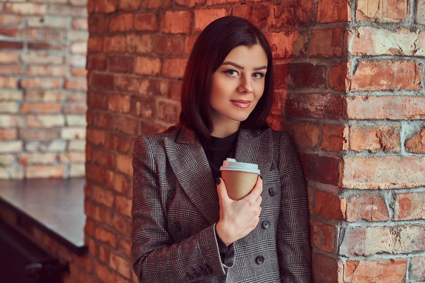 Close-up retrato de uma mulher sensual morena vestida com uma jaqueta elegante cinza segura xícara de café takeaway enquanto se inclina em uma parede de tijolo em uma sala com interior do sótão
. - Foto, Imagem