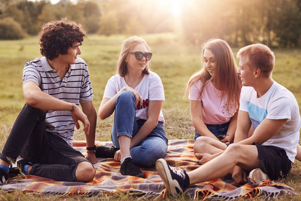 Outdoor shot of happy four friends have fun together during summer picnic, dicuss latest news, share their plans during vacation, sit on plaid on ground, have friendly relationship. Teenagers outside - Foto, imagen