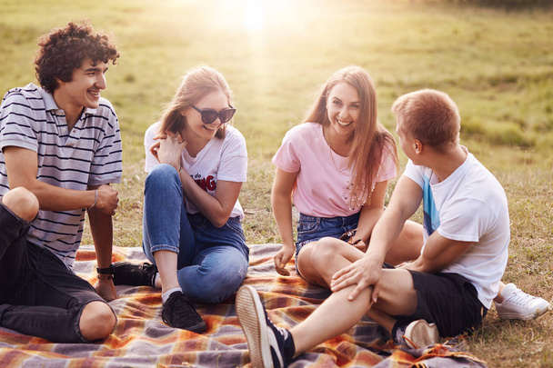 Friends, happiness and leisure concept. Photo of friendly teenagers meet together on nature, have picnic, tell each other funny stories, enjoy summer weather and sunshine, have positive smiles - 写真・画像