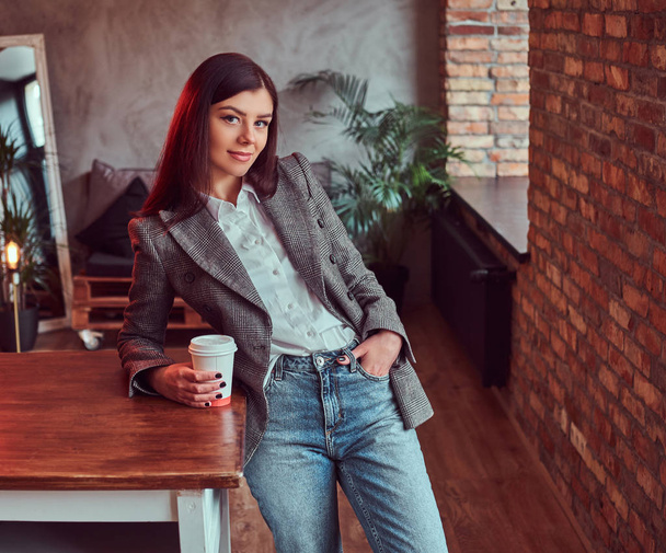 Portrait d'une jeune femme vêtue d'une élégante veste grise tenant une tasse de café à emporter tout en s'appuyant sur une table dans une chambre avec mezzanine intérieure, regardant la caméra
. - Photo, image