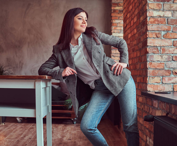 Портрет молодой женщины, одетой в серую элегантную куртку, позирующую, опираясь на стол в комнате с мансардным интерьером, отводящей взгляд
. - Фото, изображение