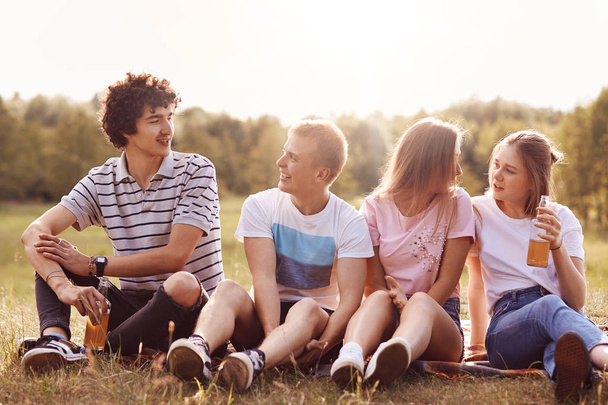 Horizontal shot of happy best four friends have pleasant talk with each other, drink cold beverage, have positive expressions, sit on grass, tell latest news, share impressions after excursion - Foto, Imagem