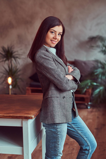 Retrato de una joven morena feliz vestida con una elegante chaqueta gris posando mientras se apoya sobre una mesa en una habitación con interior tipo loft, mirando a la cámara
. - Foto, imagen