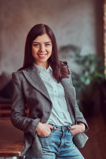 Happy young brunette woman dressed in a gray elegant jacket posing with hands in pockets while leaning on a table in a room with loft interior, looking at camera. - Фото, зображення