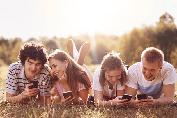 Two couples watch something in their smart phones, use internet for downloading music or watchng video, has pleased expressions, lie on ground, spend free time on nature. Friendship, lifestyle - Zdjęcie, obraz