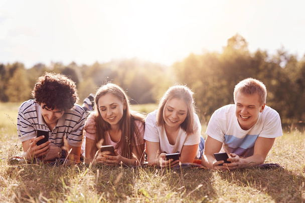 Teenagers and vacation concept. Happy youngsters lie on green grass, have positive smiles, use modern smart phones for interacting with friends and searching social networks, spend free time outdoor - Fotografie, Obrázek