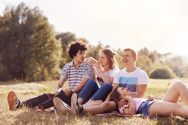 Outdoor shot of happy youngsters have charming smiles, look into distance, sit on plaid, pose against green nature and sunshine background. Four friends have good realtionship, pincic outside - Photo, Image