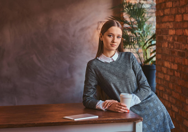 Mladá dívka oblečená v elegantní šedé šaty drží s sebou kávu a opřel se o stůl v místnosti s interiér podkroví při pohledu na fotoaparát. - Fotografie, Obrázek
