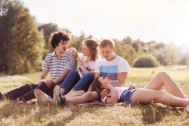 People, lifestyle and friends concept. Joyful two couples dressed in summer casual clothes, spend free time in countryside, enjoy nice weather, sit on plaid, have closeness and truthful friendship - 写真・画像