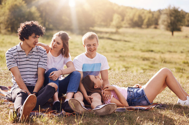 Lovely teenagers in love express mutual feeling, have positive expressions, picnic outdoor, sit on plaid, enjoys sun and green grass. Happy youngsters going to have outside activities, rest on field - Foto, Bild