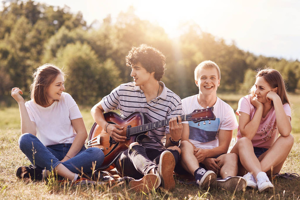 Happy young friends sing songs to guitar, have joy together, recreat outdoor, sit on green grass. Curly handsome male teenager plays guitar, enteratins his companions, enjoys summer warm day. - Φωτογραφία, εικόνα