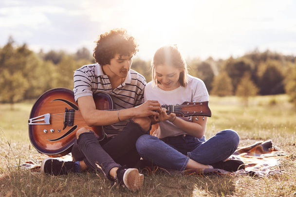 Lovely couple sit cross legs on green grass, play guitar, sing songs, enjoy togetherness. Cute young female learns how produce sound on music instrument, recieves pieces of advice from boyfriend - Foto, Imagem