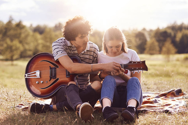 Picnic time. Lovely couple in love date on nature background, handsome male with curly hair teaches his beautiful girlfriend to play guitar, have good relationship. Dating, romance, lifestyle concept - Foto, Imagem