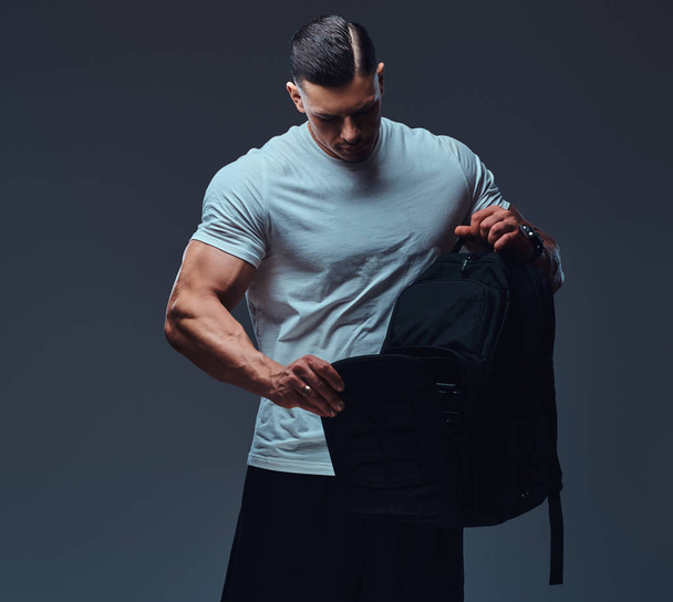 Muscular handsome bodybuilder in sportswear with rucksack looks into an open backpack in a studio. Isolated on a gray background. - Fotoğraf, Görsel