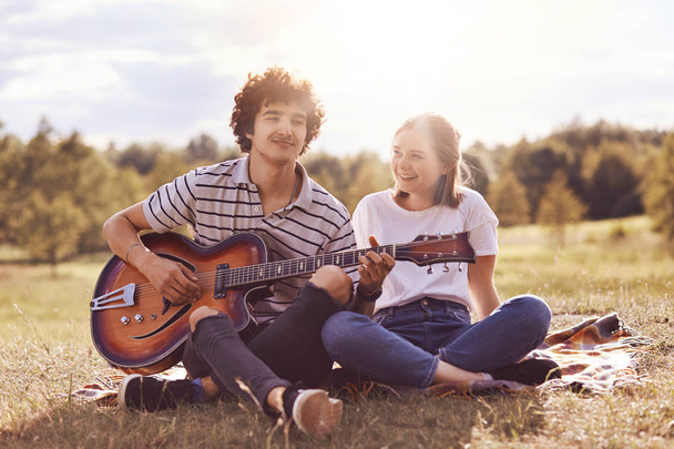 Outdoor shot of happy female and male friends sing songs and play guitar, enjoy spare time, have picnic together, smile positively, pose against blurred nature background. People and lifestyle concept - Photo, image
