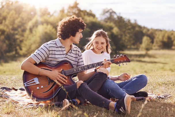 Photo of happy friends entretain themselves, sing songs and play guitar, have joyful expressions, sit on ground, dressed in casual clothes, enjoy summer sunny day on nature. Lifestyle concept - Foto, Bild