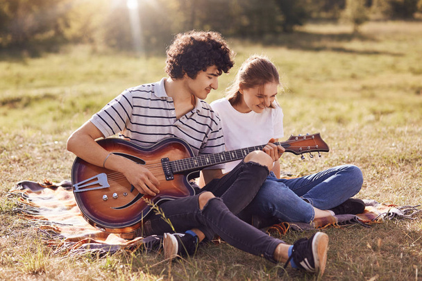 Joyful young couple have fun togehter, sing songs and play guitar, sit on plaid, have happy expressions. Attractive curly male youngster plays music instrument and entertains his girlfriend. - Fotografie, Obrázek