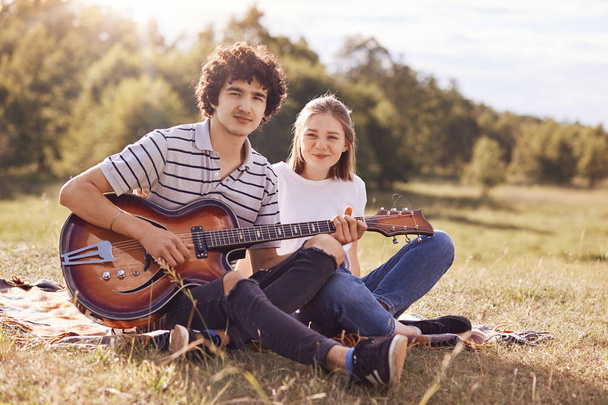 Youth, teenagers and entertainement concept. Handsome male plays guitar and sings songs to his girlfriend, look happily at camera, have joyful expressions, enjoy togetherness and beautiful nature - Photo, Image