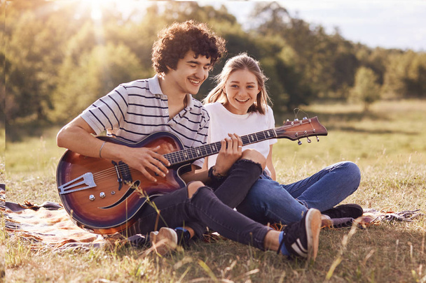 Youth, dating and recreation time concept. Happy girlfriend and boyfriend sit on green grass, play guitar, enjoy calm atmosphere, have smiles on faces. Lovely young female listens romantic songs - Photo, Image