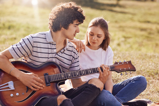 Outdoor view of lovely female and her male companion have leisure time, handsome teenager plays guitar, looks with love at his girlfriend, pose on green field. People, leisure, lifestyle concept - Foto, afbeelding